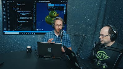 Two men with earphones, microphones,  and laptops sitting around a table and with a large screen and a starry background behind them.